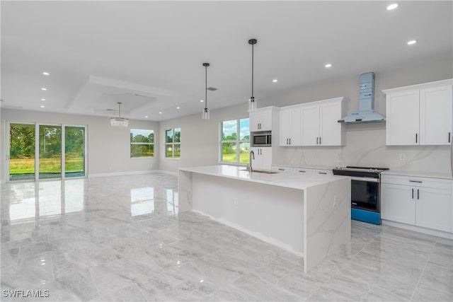 kitchen with white cabinets, appliances with stainless steel finishes, wall chimney range hood, and an island with sink