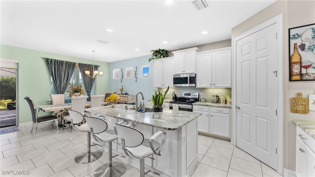 kitchen featuring appliances with stainless steel finishes, light stone countertops, a kitchen island with sink, white cabinets, and pendant lighting