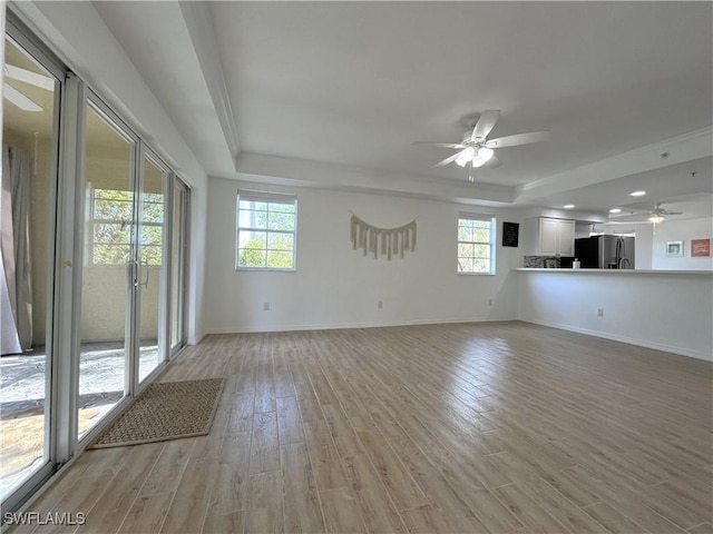 unfurnished living room with a ceiling fan, a tray ceiling, light wood-style flooring, and baseboards