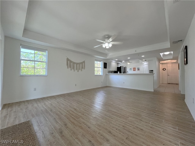 unfurnished living room featuring ceiling fan, a healthy amount of sunlight, a raised ceiling, and light hardwood / wood-style flooring