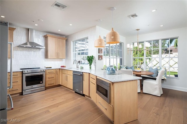 kitchen with appliances with stainless steel finishes, light brown cabinetry, decorative light fixtures, kitchen peninsula, and wall chimney range hood