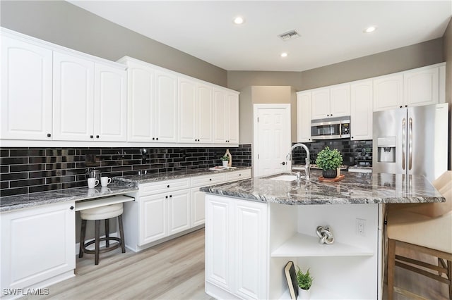 kitchen with white cabinets, stainless steel appliances, dark stone counters, and an island with sink