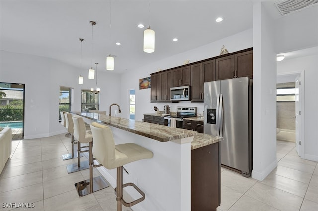 kitchen with an island with sink, appliances with stainless steel finishes, light stone counters, and pendant lighting