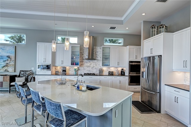 kitchen with sink, stainless steel appliances, wall chimney range hood, a raised ceiling, and an island with sink