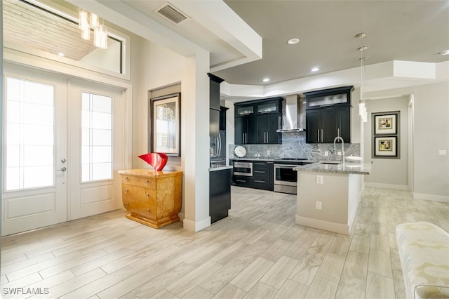 kitchen featuring wall chimney range hood, pendant lighting, a kitchen island with sink, appliances with stainless steel finishes, and light wood-type flooring