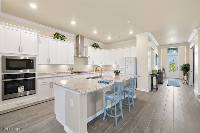 kitchen featuring light stone counters, stainless steel appliances, wall chimney range hood, white cabinetry, and an island with sink