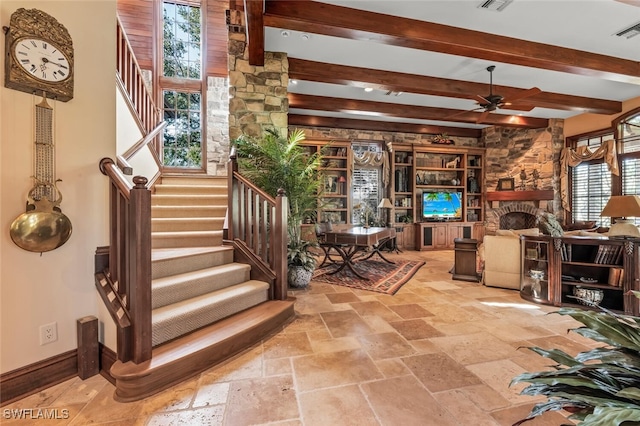 interior space with beamed ceiling, ceiling fan, a stone fireplace, and a wealth of natural light