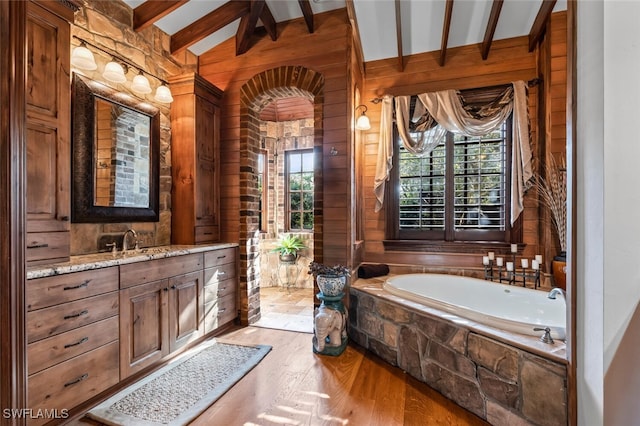 bathroom with lofted ceiling with beams, tiled bath, wood-type flooring, wooden walls, and vanity