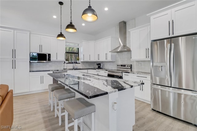 kitchen featuring white cabinets, wall chimney exhaust hood, decorative light fixtures, a kitchen island, and stainless steel appliances