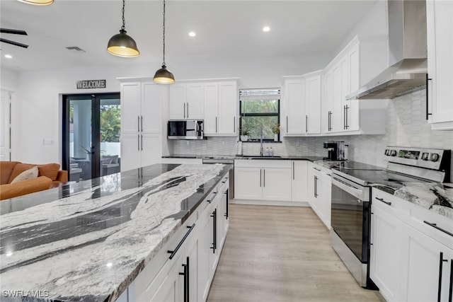 kitchen with appliances with stainless steel finishes, light stone counters, wall chimney range hood, white cabinetry, and hanging light fixtures