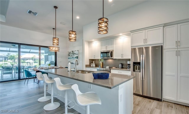 kitchen with hanging light fixtures, stainless steel appliances, light hardwood / wood-style flooring, dark stone counters, and a center island with sink