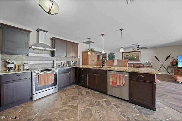 kitchen with pendant lighting, wall chimney exhaust hood, ceiling fan, appliances with stainless steel finishes, and kitchen peninsula