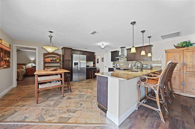 kitchen with kitchen peninsula, stainless steel fridge, decorative backsplash, dark brown cabinetry, and wall chimney range hood