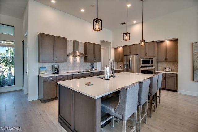 kitchen featuring a center island with sink, wall chimney exhaust hood, a towering ceiling, appliances with stainless steel finishes, and decorative light fixtures