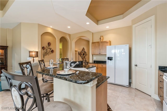 kitchen featuring sink, a raised ceiling, white refrigerator with ice dispenser, dark stone counters, and a center island with sink