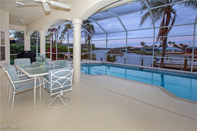 pool at dusk with ceiling fan, glass enclosure, a water view, and a patio