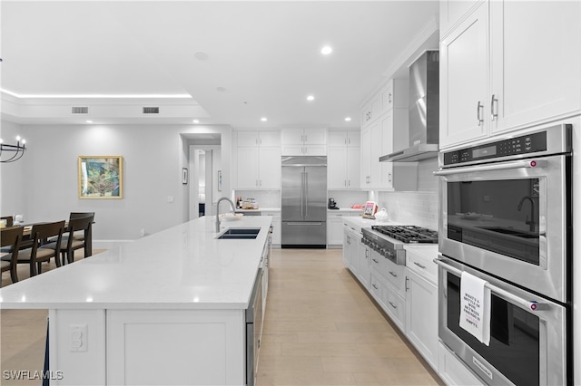 kitchen with sink, wall chimney exhaust hood, stainless steel appliances, a large island with sink, and white cabinets