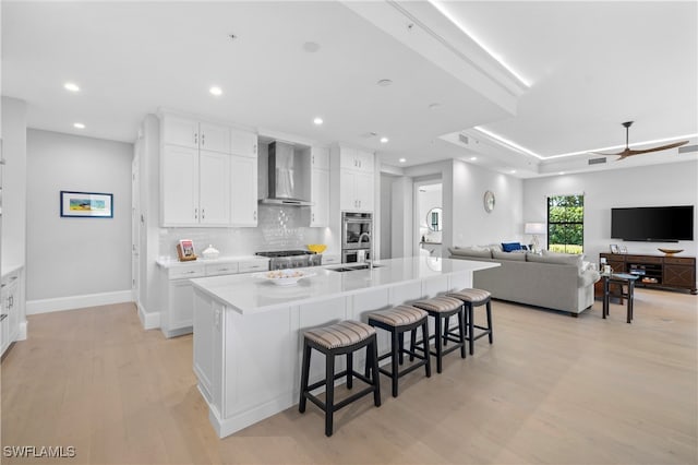 kitchen featuring light wood-type flooring, a spacious island, wall chimney range hood, white cabinetry, and a breakfast bar area