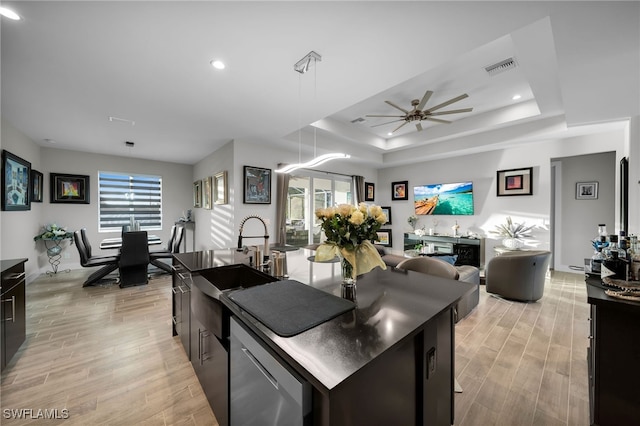 kitchen with stainless steel dishwasher, a raised ceiling, ceiling fan, light hardwood / wood-style floors, and an island with sink