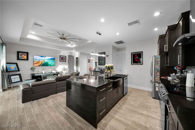 kitchen with a raised ceiling, an island with sink, stainless steel appliances, and light wood-type flooring