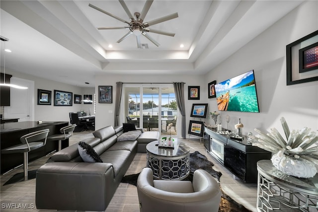 living room with a raised ceiling, bar, ceiling fan, and light wood-type flooring