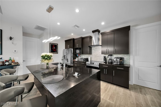 kitchen featuring a center island with sink, wall chimney exhaust hood, light wood-type flooring, tasteful backsplash, and decorative light fixtures