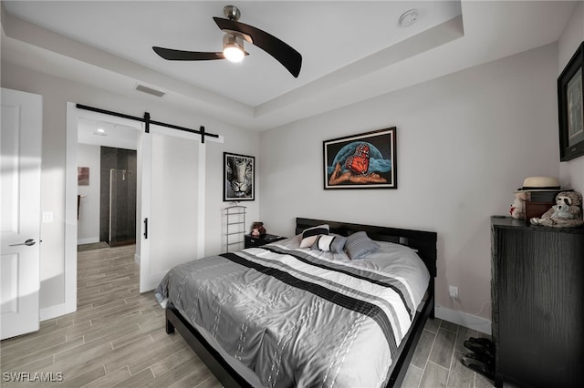 bedroom featuring a barn door, a tray ceiling, light hardwood / wood-style flooring, and ceiling fan