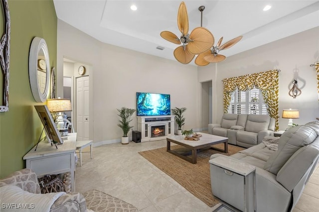 living area with a raised ceiling, visible vents, ceiling fan, a lit fireplace, and baseboards