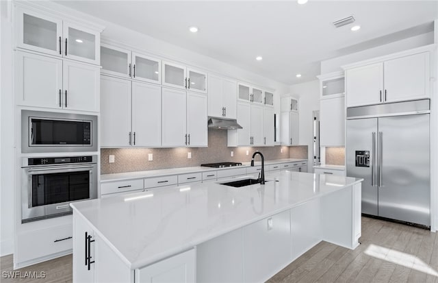 kitchen with built in appliances, a center island with sink, white cabinetry, and sink