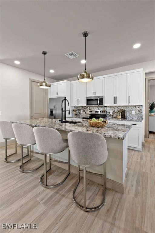 kitchen with light stone countertops, stainless steel appliances, light hardwood / wood-style flooring, pendant lighting, and a kitchen island with sink