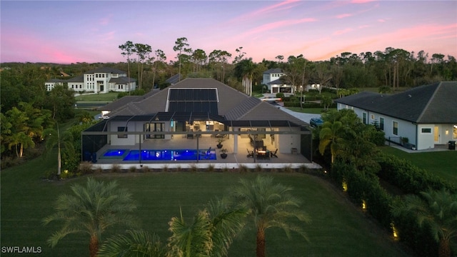 back house at dusk featuring a lawn, glass enclosure, a patio area, and solar panels