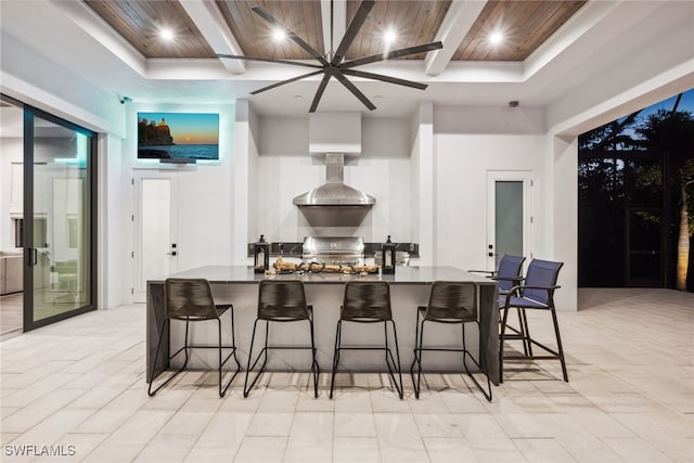 kitchen featuring a kitchen bar, a tray ceiling, wall chimney exhaust hood, and wood ceiling