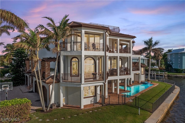 back house at dusk with a lawn, a balcony, and a fenced in pool