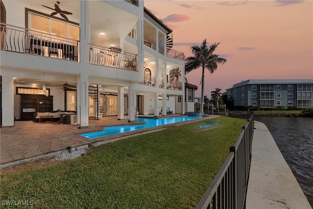 back house at dusk with a fenced in pool, ceiling fan, a balcony, a yard, and a patio area