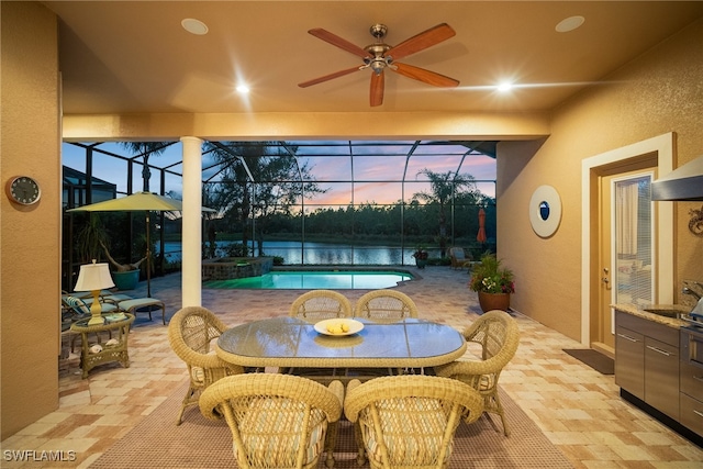 patio terrace at dusk featuring glass enclosure, ceiling fan, and a water view