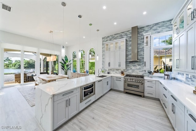 kitchen featuring stainless steel appliances, wall chimney range hood, light stone counters, backsplash, and pendant lighting