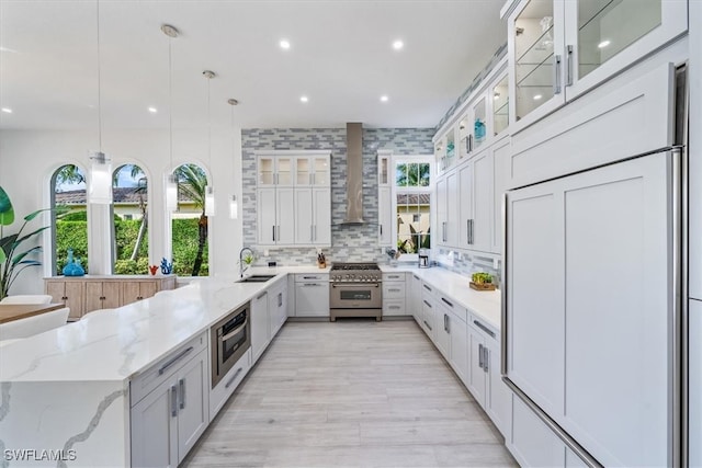 kitchen with white cabinets, wall chimney range hood, sink, hanging light fixtures, and appliances with stainless steel finishes