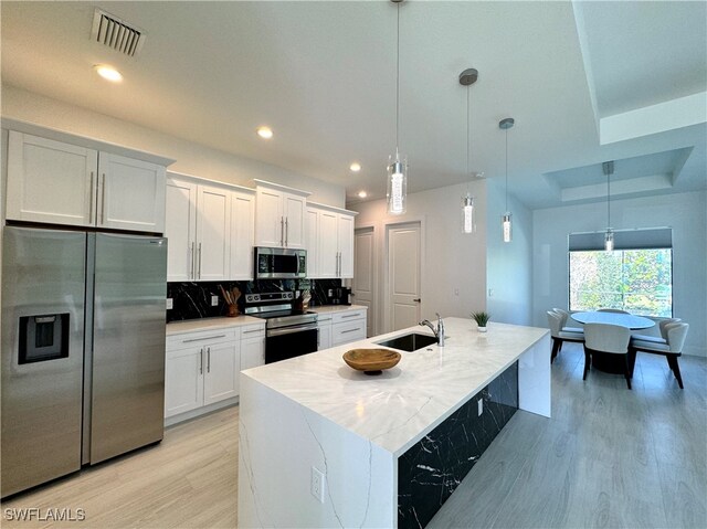 kitchen featuring decorative backsplash, appliances with stainless steel finishes, white cabinets, hanging light fixtures, and an island with sink