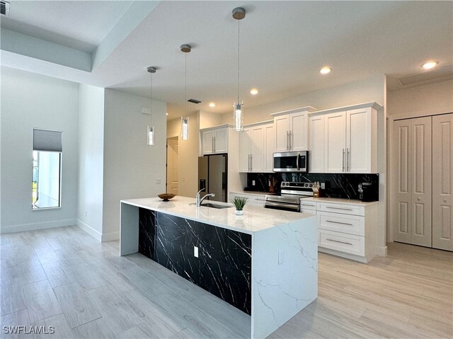 kitchen with light wood-type flooring, stainless steel appliances, a kitchen island with sink, white cabinets, and hanging light fixtures