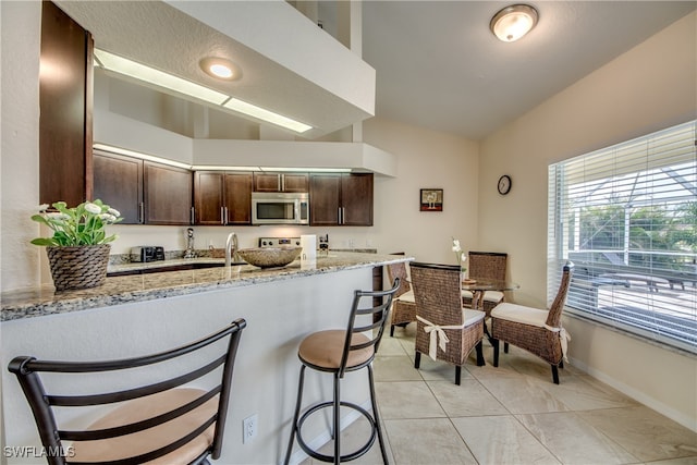 kitchen with dark brown cabinetry, a kitchen breakfast bar, light stone counters, kitchen peninsula, and vaulted ceiling