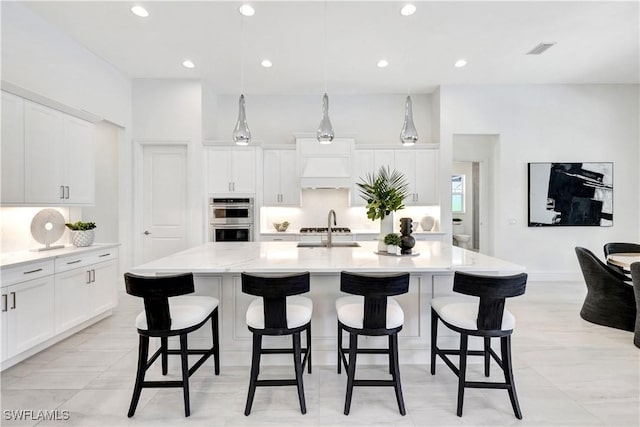 kitchen featuring pendant lighting, white cabinets, stainless steel double oven, and a kitchen island with sink