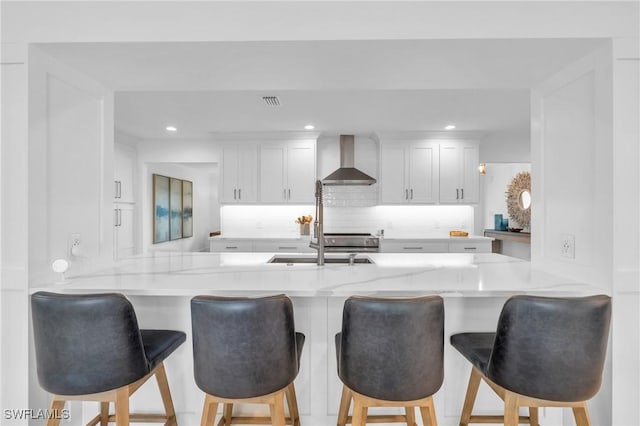 kitchen with kitchen peninsula, a kitchen bar, backsplash, wall chimney range hood, and white cabinets