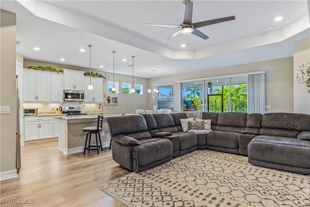 living room with ceiling fan, recessed lighting, baseboards, light wood finished floors, and a tray ceiling