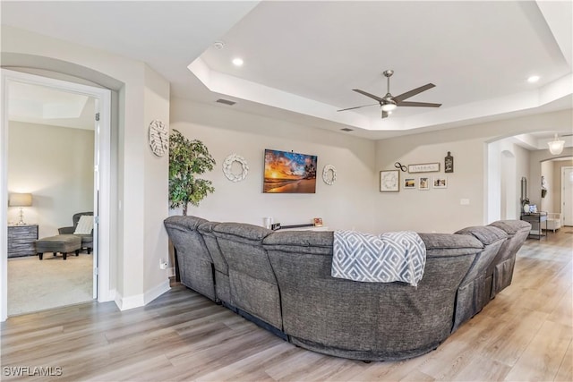 living area featuring light wood-style floors, ceiling fan, a tray ceiling, and arched walkways