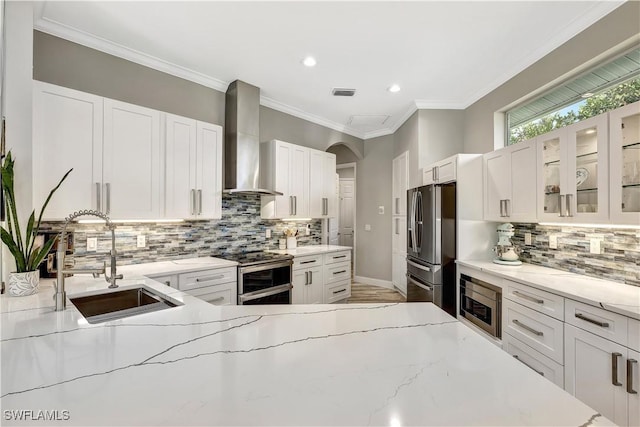 kitchen with appliances with stainless steel finishes, glass insert cabinets, white cabinetry, a sink, and wall chimney exhaust hood