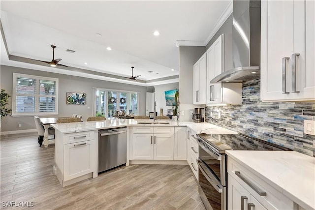 kitchen with white cabinets, appliances with stainless steel finishes, a peninsula, a tray ceiling, and wall chimney range hood