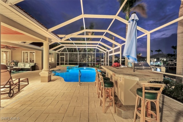 pool at dusk featuring area for grilling, glass enclosure, an outdoor bar, and a patio area