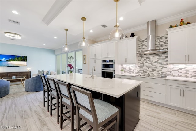 kitchen with light stone countertops, a kitchen island with sink, wall chimney range hood, pendant lighting, and white cabinetry