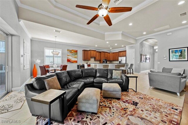 living room with ceiling fan with notable chandelier, a raised ceiling, crown molding, and light tile patterned flooring