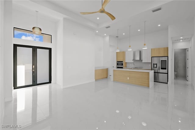 kitchen with hanging light fixtures, wall chimney range hood, a spacious island, light brown cabinetry, and appliances with stainless steel finishes